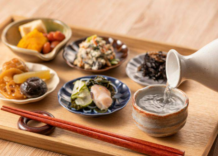 A serving tray of different Japanese small plates. In the corner, someone is pouring sake.